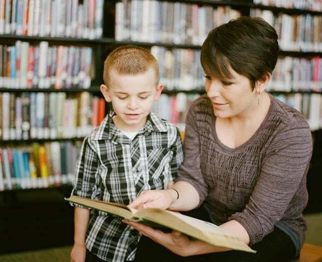 Mother Teaching Boy Child. Boy in gray sweater beside boy in gray and white plaid dress shirt.